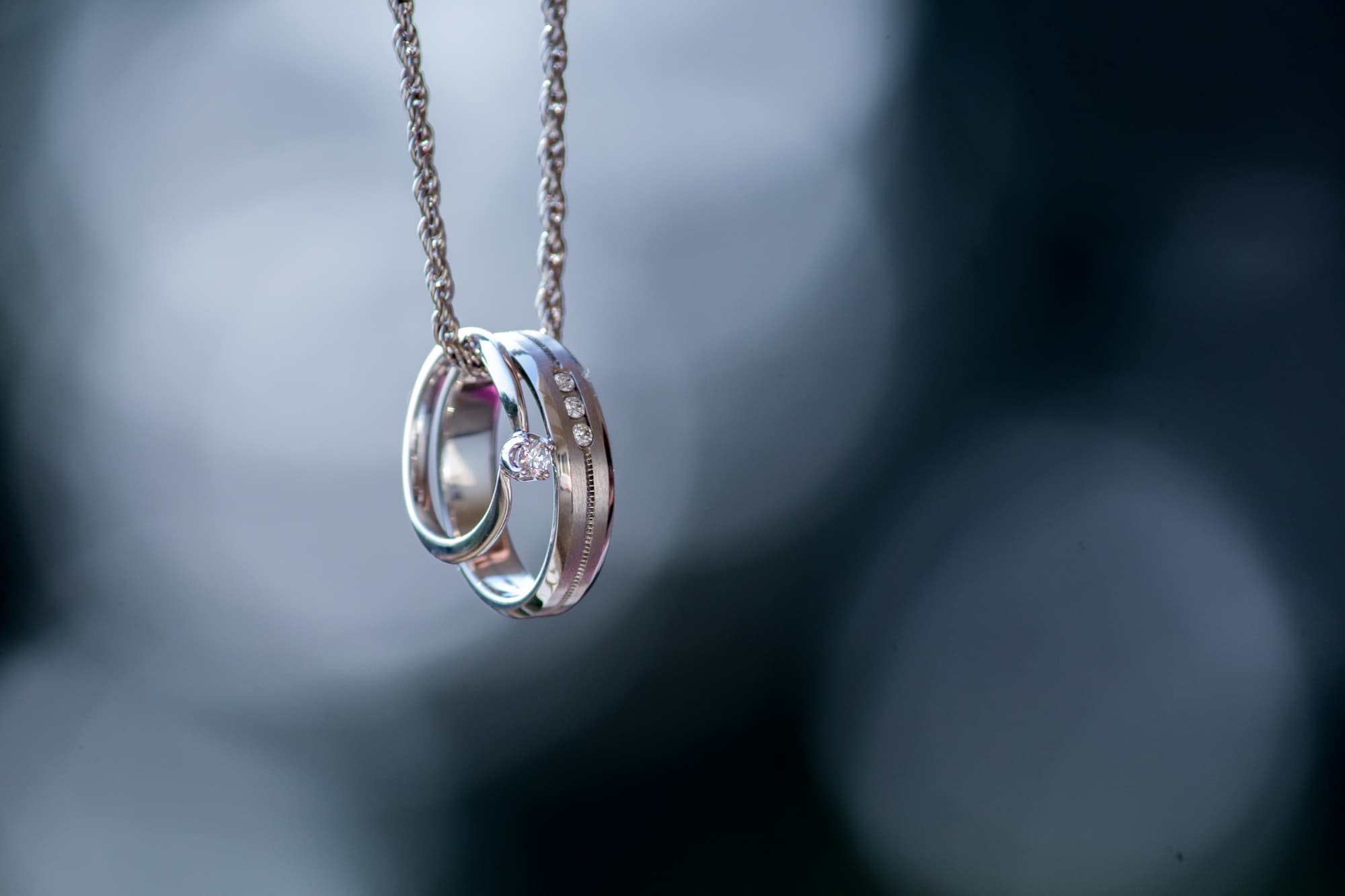 Engagement and wedding bands hanging from a silver chain against dark bokeh background.